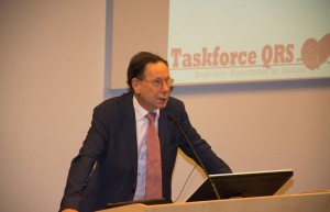 Luc Soete staat achter een microfoon en achter hem is het logo van Taskforce QRS op een presentatiescherm te zien