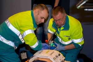 Twee ambulancemensen zitten bij een reanimatiepop en geven beademingen met een ballon en masker. De AutoPulse is aangesloten voor borstcompressies.