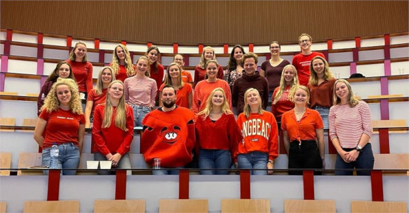 23 instructeurs staan tussen de banken van een collegezaal, hebben allemaal rode kleding aan en één instructeur draagt een kostuum van een hart